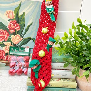 Puede incluir: Calcetín de Navidad rojo de crochet con un lazo en la parte superior y tres pequeños adornos de duendes colgando. El calcetín está decorado con campanas verdes y amarillas.