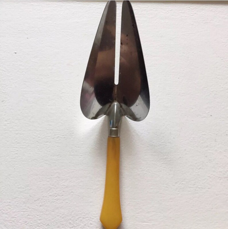 May include: A vintage cake server with a yellow handle and a silver, two-pronged blade. The server is designed to cut and serve cake.
