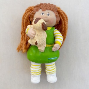 Puede incluir: Una muñeca vintage de Cabbage Patch Kids con cabello de hilo rojo, que lleva un vestido verde y mangas y leggings a rayas amarillas y blancas. La muñeca sostiene un pequeño perro beige con manchas marrones. La cara de la muñeca tiene mejillas rosadas y ojos azules.