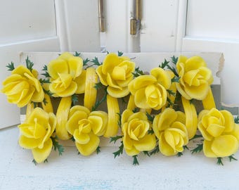 May include: A collection of bright yellow artificial roses with green leaves and yellow ribbons. The roses are arranged on a vintage card, creating a cheerful floral display. The roses are a vibrant yellow color.