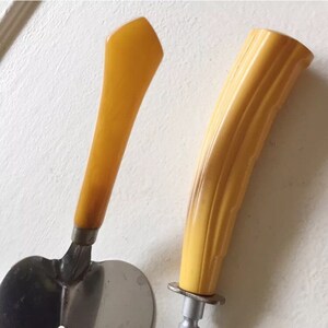 May include: Two vintage butter knives with yellow plastic handles. One knife has a rounded handle and the other has a pointed handle. Both knives have metal blades.
