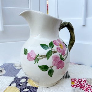 Puede incluir: Jarra de cerámica color crema con diseño floral. La jarra presenta flores rosas, hojas verdes y un asa marrón en forma de rama. Las flores tienen centros amarillos. La jarra se utiliza probablemente para servir bebidas.