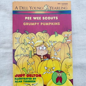 Puede incluir: Un libro de bolsillo Dell Young Yearling titulado "Pee Wee Scouts Grumpy Pumpkins" de Judy Delton, ilustrado por Alan Tiegreen. La portada presenta un grupo de calabazas naranjas con caras enojadas y una niña en el centro.