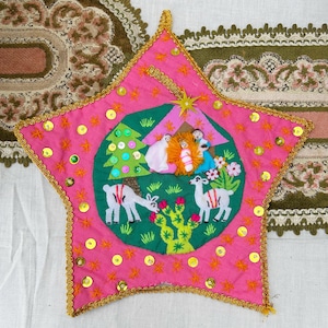 Può includere: Un ornamento a forma di stella rosa con un bordo dorato, che presenta una scena della natività ricamata con due lama e un albero di Natale. L'ornamento è decorato con paillettes e stelle.