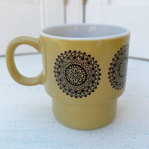 Könnte beinhalten: Eine gelbe Keramiktasse mit einem schwarz-weißen geometrischen Muster. Die Tasse hat einen breiten, abgerundeten Griff.