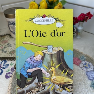 May include: A vintage children's book titled "L'Oie d'or" with a yellow cover. The cover illustration depicts a young man kneeling before a golden goose, with an axe on a tree stump. The book has the "Coccinelle" logo.