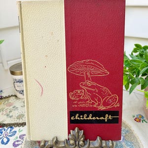 Könnte beinhalten: Vintage "Childcraft" Buch mit cremefarbenem Buchrücken und rotem Einband mit einer goldenen Illustration eines Pilzes und Fröschen. Das Wort "childcraft" ist goldfarben auf einem schwarzen Rechteck gedruckt. Das Buch wird in einem dekorativen Metallständer präsentiert.