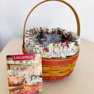 May include: A Longaberger 1998 Mother's Day basket with a wooden handle and a floral fabric liner. The basket is woven with yellow and red accents. A brochure with the text "Mother's Day Basket" is next to the basket.