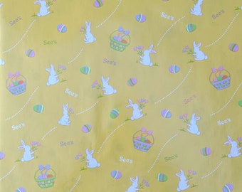 Pode incluir: Papel de embrulho amarelo com um padrão repetido de coelhos brancos segurando flores cor-de-rosa, ovos de Páscoa pastel e cestas de ovos de Páscoa. O texto "See's" é repetido em todo o padrão.