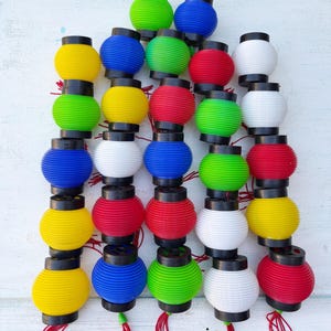 May include: A collection of colorful, small paper lanterns in various colors, including yellow, blue, green, red, and white. Each lantern has a black top and bottom, and some have red tassels. The lanterns are stacked together.