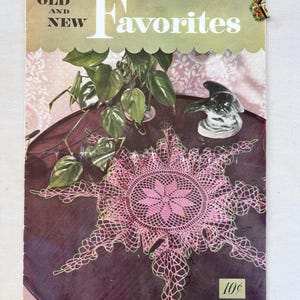 以下が含まれることがあります： 「Old and New Favorites」と題されたヴィンテージ本の表紙。ピンク色のレース編みのドイリーと小さな鳥の置物が、濃い木製のテーブルの上に置かれています。表紙には「CLARK'S ONT J&P COATS」と「BOOK No. 269」の文字があります。緑色の植物も見えます。