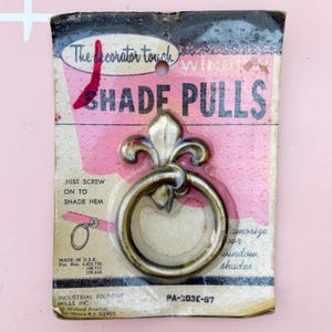 Op de afbeelding: Vintage raamgordijn trekt aan originele verpakking. De trekkoorden zijn gemaakt van messing en hebben een fleur-de-lis ontwerp. De verpakking heeft de tekst "SHADE PULLS" en "The decorator touch".