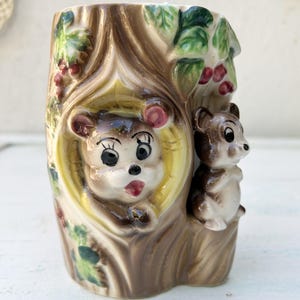 Peut inclure: Un vase vintage en céramique en forme de tronc d'arbre, avec deux ours de dessin animé. L'un est à l'intérieur d'une ouverture à bord jaune, l'autre est à l'extérieur. Le vase est peint dans des tons de marron, beige et vert, avec des accents de baies rouges.