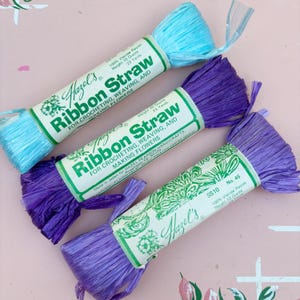 Puede incluir: Tres paquetes de cinta de paja Hazel's, uno azul claro y dos morados, cada uno envuelto en una etiqueta de papel blanco con texto verde. Las etiquetas indican 100% rayón de viscosa, 10 gramos y 23 yardas para ganchillo, tejido y flores.