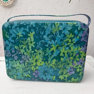 Könnte beinhalten: Eine vintage Handtasche in Türkis mit einem Blumenmuster in Blau-, Grün- und Lilatönen. Die Tasche hat einen oberen Griff und einen Reißverschluss. Die Gesamtform ist rechteckig mit abgerundeten Ecken.