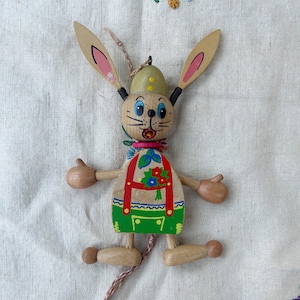 May include: Wooden bunny figurine with articulated limbs, wearing a green and beige outfit with red braces and floral accents. The bunny has long ears with pink interiors, blue eyes, and a painted expression. A string is attached for hanging.