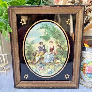 May include: A framed oval-shaped print of a couple sitting in a garden setting. The man is wearing a blue jacket and the woman is wearing a red dress. The print is surrounded by a black background with gold stars.