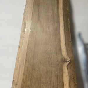 May include: A long, narrow wooden slab with a natural, rustic finish. The wood has a light brown color with darker brown streaks and knots. The slab has a live edge on one side, giving it a unique and organic look.