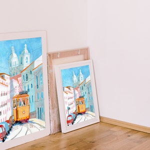 May include: A watercolor painting of a yellow tram traveling up a hill in a city with white and blue buildings. The painting is framed in a white frame and is leaning against a white wall.