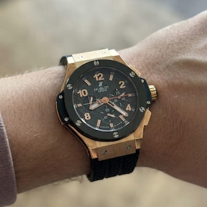 Op de afbeelding: Een Hublot Geneve chronograaf polshorloge met een goudkleurige kast, een zwarte ring vastgezet met zes schroeven en een zwarte wijzerplaat met goudkleurige cijfers en wijzers. Het horloge heeft een zwarte, gestructureerde rubberen horlogeband.
