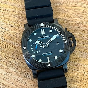May include: A Panerai Submersible automatic watch with a black dial, luminous markers, and a date window at the three o'clock position. The watch features a black textured bezel, a black rubber strap, and text reading Panerai Submersible Automatic 300m/1000ft.