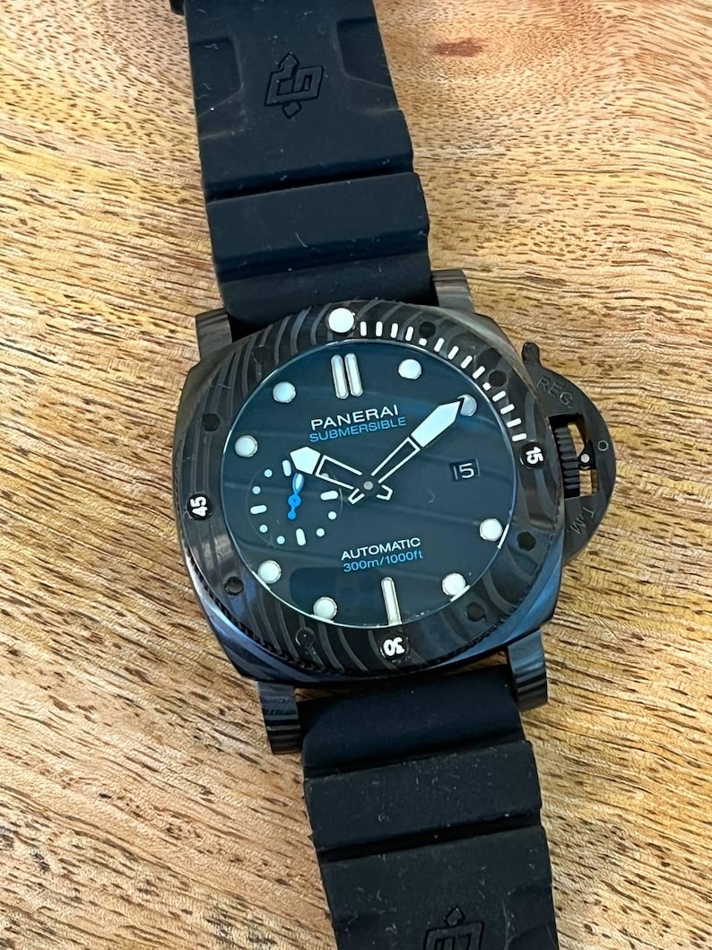 May include: A Panerai Submersible automatic watch with a black dial, luminous markers, and a date window at the three o'clock position. The watch features a black textured bezel, a black rubber strap, and text reading Panerai Submersible Automatic 300m/1000ft.