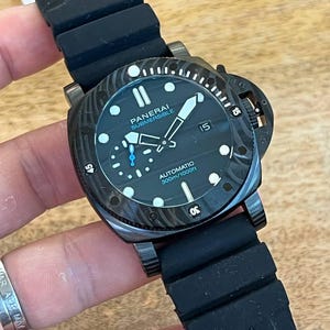 May include: A black Panerai Submersible automatic watch with a textured case, black rubber strap, and a black dial featuring luminous markers, a date window at the three o'clock position, and text reading Panerai Submersible Automatic 300m/1000ft.