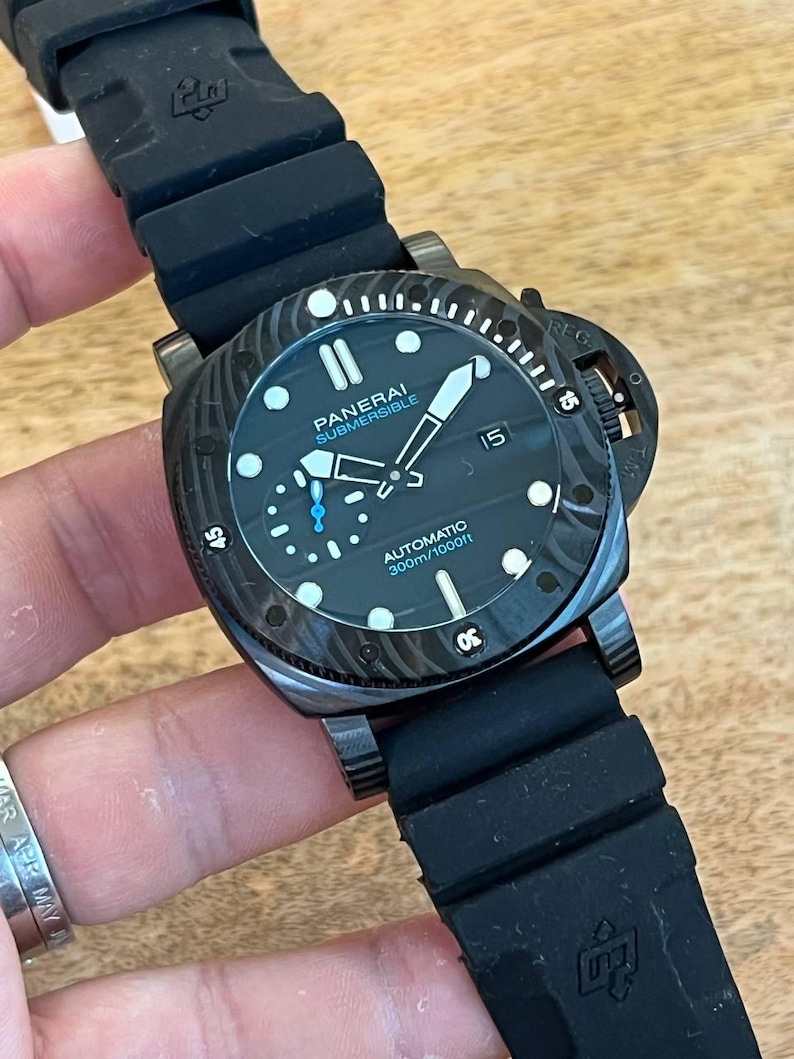 May include: A black Panerai Submersible automatic watch with a textured case, black rubber strap, and a black dial featuring luminous markers, a date window at the three o'clock position, and text reading Panerai Submersible Automatic 300m/1000ft.