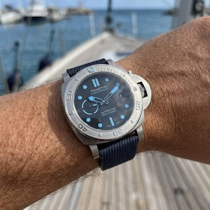May include: A Panerai Submersible automatic watch with a brushed silver-tone stainless steel case, a black dial featuring light blue luminous markers and hands, and a dark blue fabric strap with a secure buckle closure system.