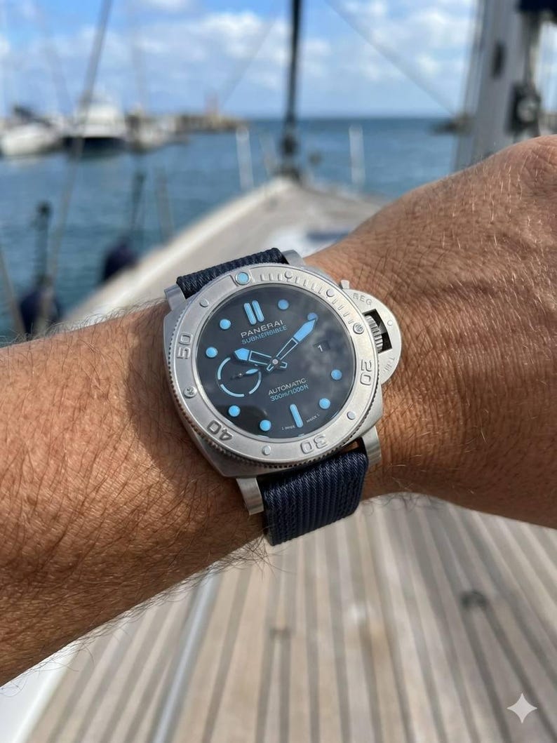 May include: A Panerai Submersible automatic watch with a brushed silver-tone stainless steel case, a black dial featuring light blue luminous markers and hands, and a dark blue fabric strap with a secure buckle closure system.