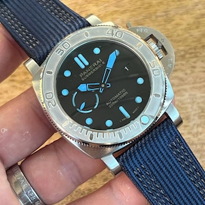 May include: A Panerai Submersible automatic watch with a black dial, light blue luminous hands and hour markers, and a silver-tone stainless steel case. The watch features a rotating bezel with engraved numerals and a dark blue textured fabric strap.