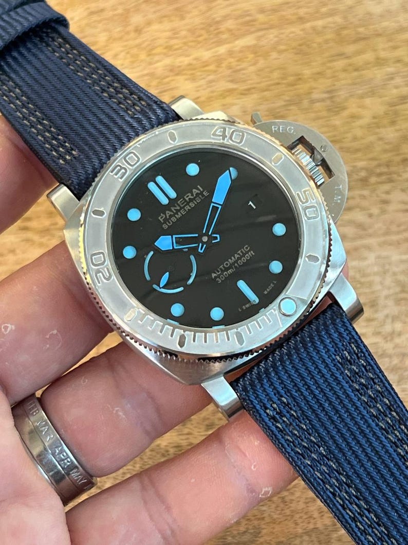 May include: A Panerai Submersible automatic watch with a black dial, light blue luminous hands and hour markers, and a silver-tone stainless steel case. The watch features a rotating bezel with engraved numerals and a dark blue textured fabric strap.