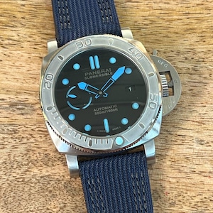 May include: A Panerai Submersible automatic watch with a black dial, luminous light blue hour markers, and hands. The watch features a silver-tone stainless steel case, a rotating bezel with engraved numerals, and a textured dark blue fabric strap.