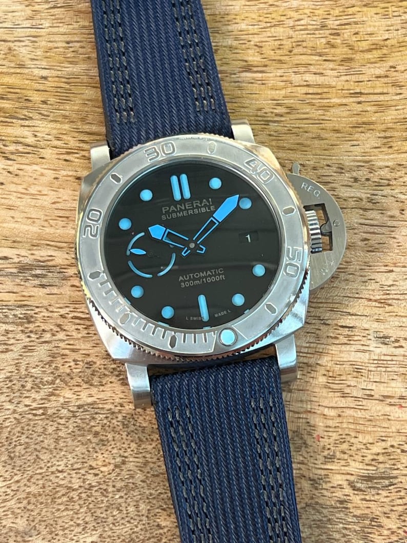 May include: A Panerai Submersible automatic watch with a black dial, luminous light blue hour markers, and hands. The watch features a silver-tone stainless steel case, a rotating bezel with engraved numerals, and a textured dark blue fabric strap.
