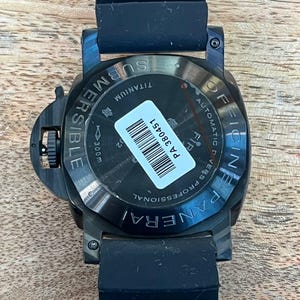 May include: A black Officine Panerai Submersible watch with a black rubber strap. The case back is made of titanium and features the text "OFFICINE PANERAI", "SUBMERSIBLE", "AUTOMATIC DIVERS PROFESSIONAL", and "300m". A white barcode sticker reads "PA 380451".