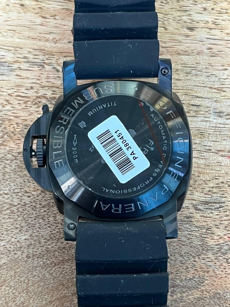 May include: A black Officine Panerai Submersible watch with a black rubber strap. The case back is made of titanium and features the text "OFFICINE PANERAI", "SUBMERSIBLE", "AUTOMATIC DIVERS PROFESSIONAL", and "300m". A white barcode sticker reads "PA 380451".