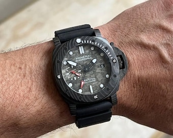 May include: A black Panerai Submersible watch with a textured gray dial, luminous hour markers, and a date window at the three o'clock position. The watch features a black rubber strap, a rotating bezel, and a crown protection bridge on the side.