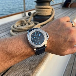 May include: A Panerai wristwatch with a round black dial, luminous hands, and hour markers, featuring a silver-tone stainless steel case and a textured dark blue fabric strap. The watch face displays the Panerai logo and automatic text.