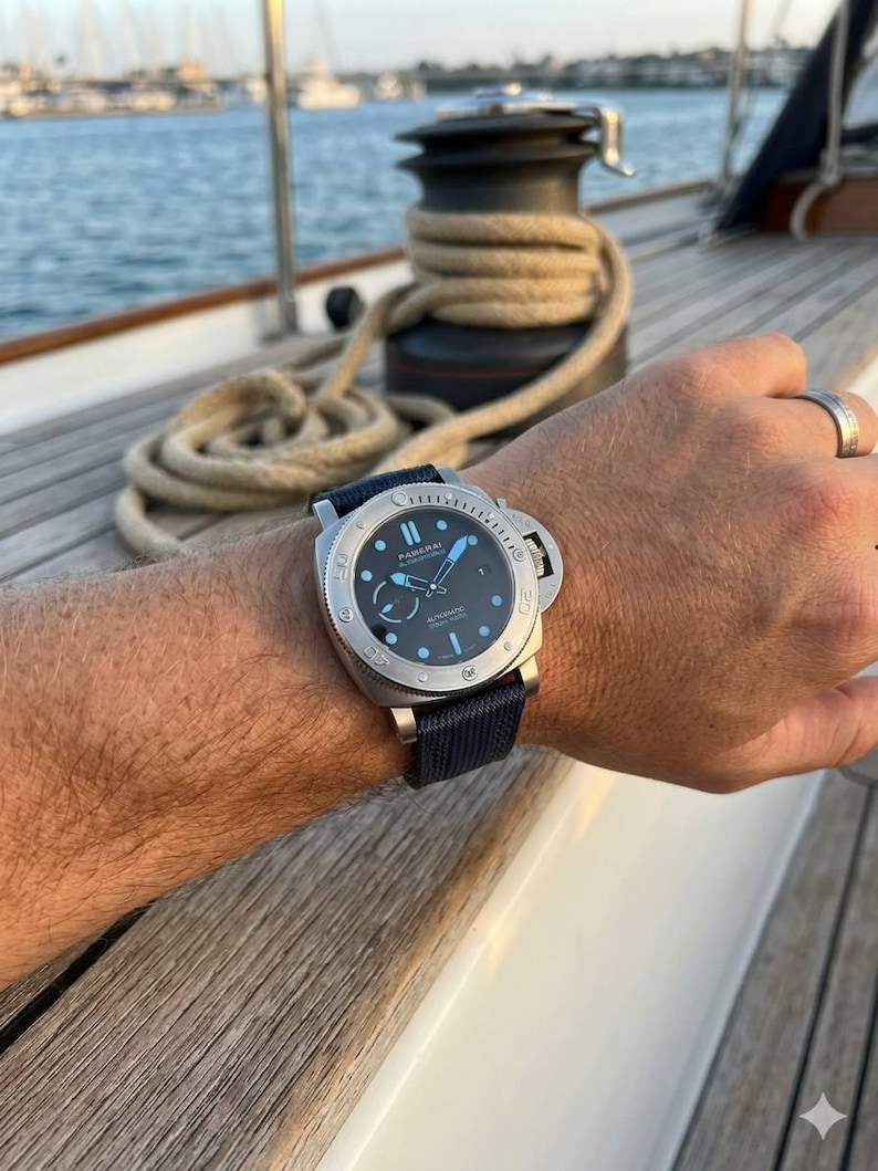 May include: A Panerai wristwatch with a round black dial, luminous hands, and hour markers, featuring a silver-tone stainless steel case and a textured dark blue fabric strap. The watch face displays the Panerai logo and automatic text.