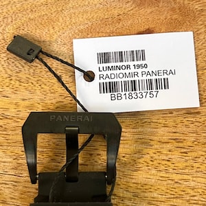 May include: A black watch buckle with the word PANERAI engraved on the top bar. A white rectangular tag is attached with a black cord, featuring the text LUMINOR 1950, RADIOMIR PANERAI, and the alphanumeric code BB1833757 with two barcodes.