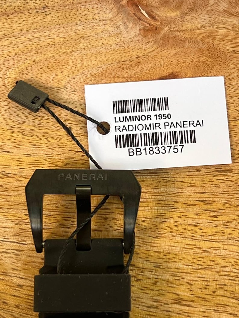 May include: A black watch buckle with the word PANERAI engraved on the top bar. A white rectangular tag is attached with a black cord, featuring the text LUMINOR 1950, RADIOMIR PANERAI, and the alphanumeric code BB1833757 with two barcodes.