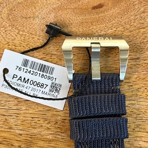May include: A silver-toned metal watch buckle engraved with the word PANERAI, attached to a dark blue textured fabric watch strap. A white rectangular tag is tied to the buckle, displaying a barcode and the text PAM00687 RADIOMIR 47 2017 MARINA.