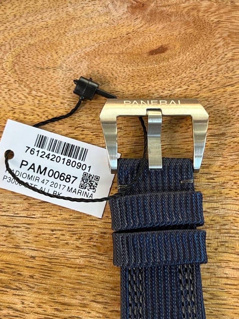 May include: A silver-toned metal watch buckle engraved with the word PANERAI, attached to a dark blue textured fabric watch strap. A white rectangular tag is tied to the buckle, displaying a barcode and the text PAM00687 RADIOMIR 47 2017 MARINA.