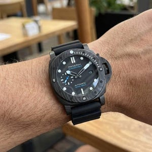 May include: A black Panerai Submersible automatic watch with a textured carbon fiber case, a black rubber strap, a rotating bezel with numerical markers, a date window at the three o'clock position, and a small seconds sub-dial at nine o'clock.