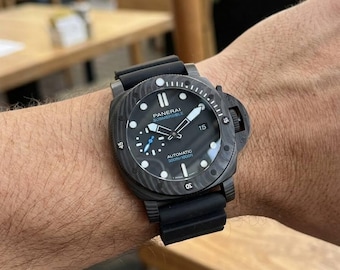 May include: A black Panerai Submersible automatic watch with a textured carbon fiber case, a black rubber strap, a rotating bezel with numerical markers, a date window at the three o'clock position, and a small seconds sub-dial at nine o'clock.