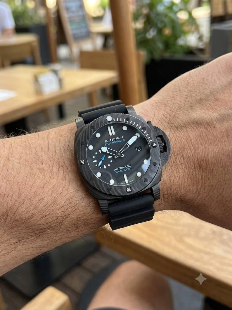 May include: A black Panerai Submersible automatic watch with a textured carbon fiber case, a black rubber strap, a rotating bezel with numerical markers, a date window at the three o'clock position, and a small seconds sub-dial at nine o'clock.