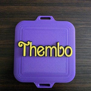 May include: A purple, rectangular item with rounded corners and handle-shaped cutouts. The word "Thembo" is in yellow, stylized lettering on the surface. This could be a decorative piece or a personalised accessory.