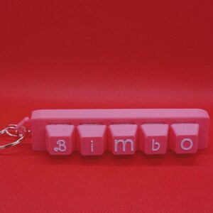 以下が含まれることがあります： 白い文字で「bimbo」と書かれたピンク色のキーボードキーのキーホルダー。キーホルダーはプラスチック製で、シルバーのキーリングが付いています。背景は無地の赤色です。