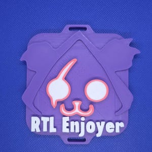 Op de afbeelding: Een paarse en oranje badge met de tekst "RTL Enjoyer". De badge heeft een gestileerd cartoongezicht met grote witte ogen en een roze mond. De badge heeft een rechthoekige vorm met een gat aan de bovenkant om aan een koord te bevestigen.