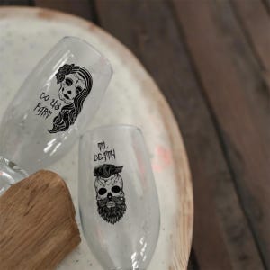 May include: Two clear glass champagne flutes with black skull and floral designs. The text on the glasses reads "Do Us Part" and "Til Death".